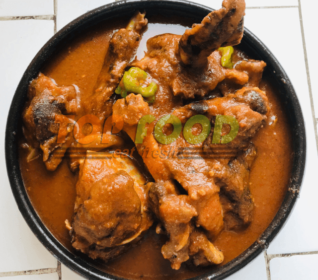 Bowl of rich red soup with assorted meats and green vegetables, served on a tiled surface.