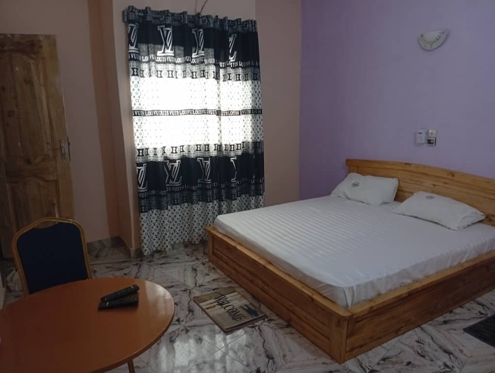 Simple bedroom with a wooden bed, white bedding, patterned curtains, and a small round table.