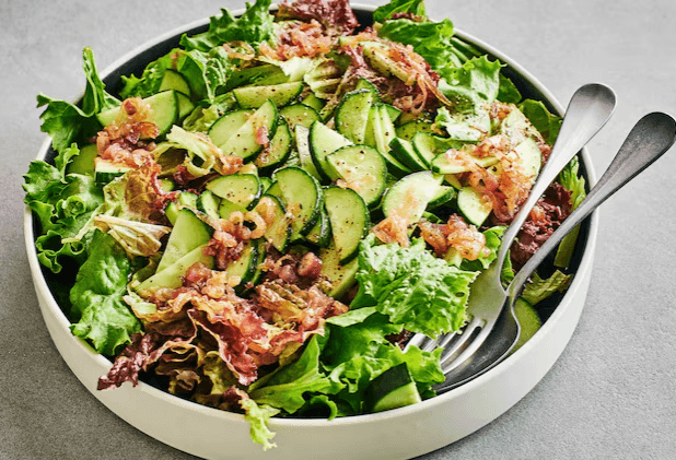 Bowl of mixed green salad with cucumbers and bacon, served with a fork and spoon.