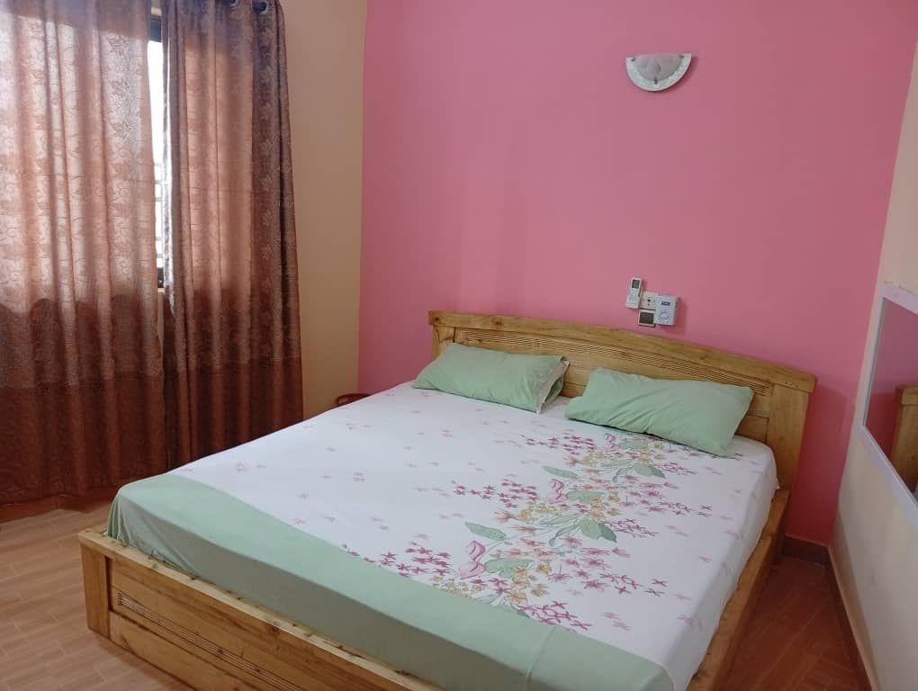 Bedroom with wooden bed, green pillows, floral bedsheet, pink wall, and brown curtains.